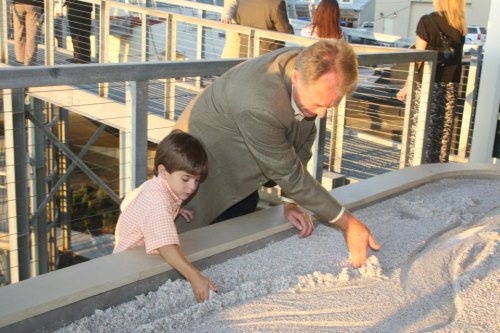 A man and a young boy play together, shaping sand in a large container on a balcony. People are visible in the background.