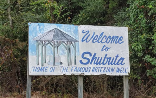 Sign welcoming visitors to Shubrita, highlighting its famous artesian well, surrounded by greenery.