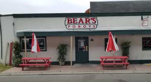 Exterior of Bear's PoBoys restaurant featuring red picnic tables and umbrellas, with a simple white and green facade.