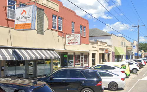 A street view featuring shops and parked cars under a blue sky with clouds.
