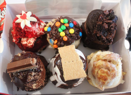 A box of six decorated cupcakes with various toppings, including chocolate, cookies, and colorful candies.