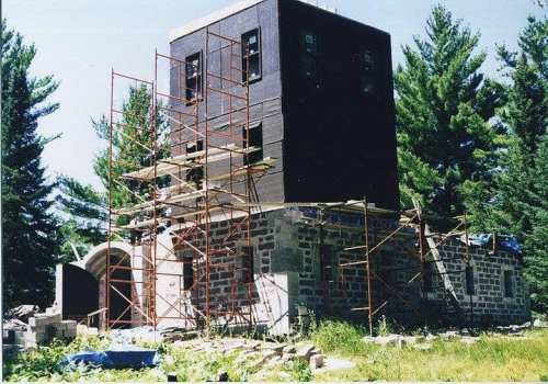 A partially constructed building with scaffolding, surrounded by trees and overgrown grass.