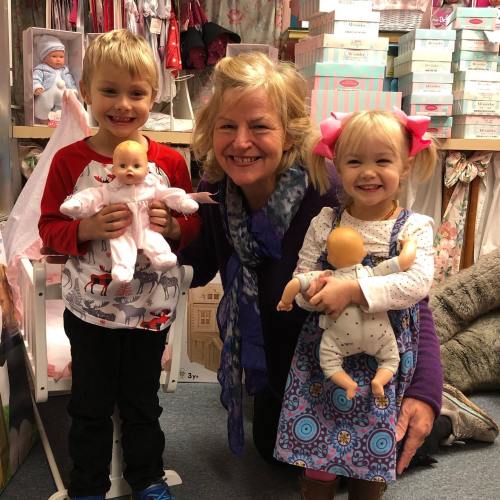 Two children hold dolls while posing with a smiling woman in a colorful store filled with toys and clothing.