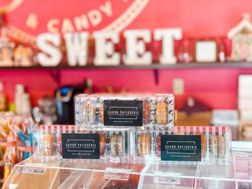 Three boxes of macarons displayed in a colorful candy shop, with a "SWEET" sign in the background.