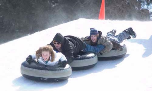 Three people on snow tubes sliding down a snowy hill, with a bright orange cone in the background.