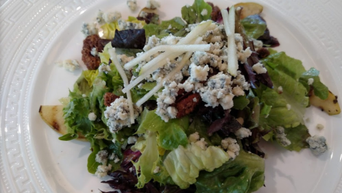 A fresh salad with mixed greens, blue cheese crumbles, sliced apples, and crunchy toppings on a white plate.