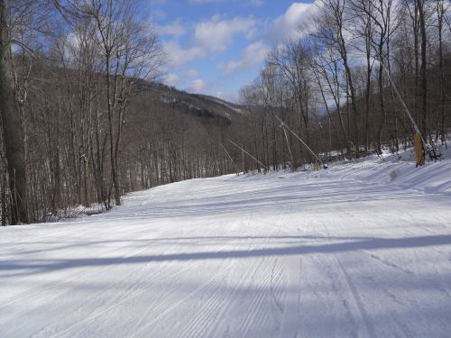 A snowy ski slope surrounded by bare trees under a blue sky with scattered clouds.