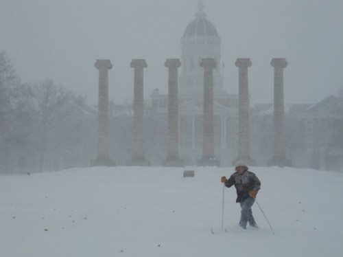 A person skis through deep snow in a blizzard, with tall stone columns and a dome building in the background.