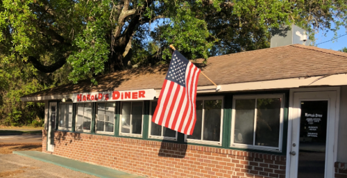 A brick diner with a sign reading "Harold's Diner" and an American flag hanging outside, surrounded by trees.