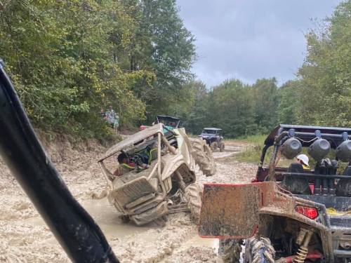 Off-road vehicles navigating a muddy trail, with one vehicle tilted and covered in mud, surrounded by trees.
