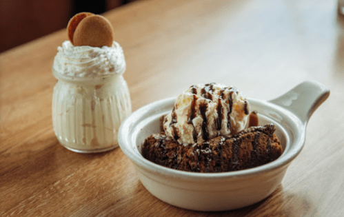 A bowl of brownie topped with ice cream and chocolate sauce, alongside a jar of whipped cream and cookies.