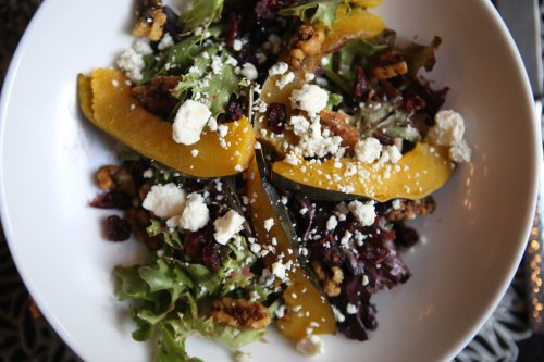 A fresh salad with mixed greens, roasted squash, walnuts, cranberries, and crumbled feta cheese in a white bowl.