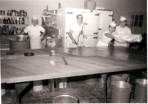 Four people in a kitchen, wearing white uniforms and hats, working around a large wooden table with various kitchen supplies.