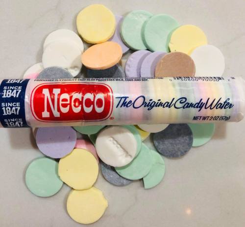 A roll of Necco candy wafers surrounded by colorful, round candy pieces on a light surface.