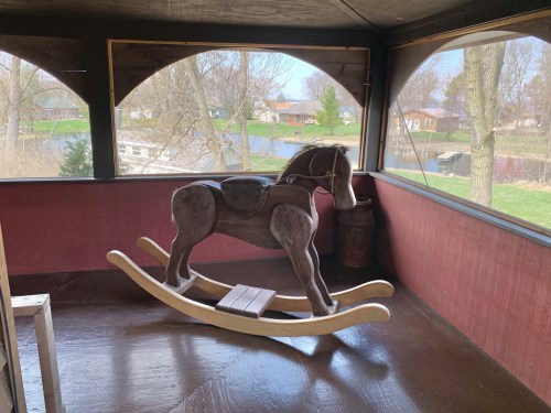 A wooden rocking horse sits on a porch with a view of a lake and trees in the background.