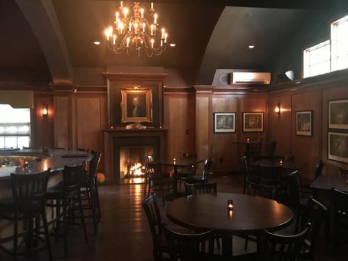 Cozy interior of a dimly lit bar with wooden walls, a fireplace, and tables set for dining.