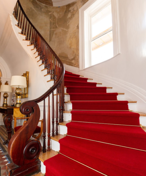 A grand staircase with a curved wooden railing and red carpet, leading to a bright, airy landing.