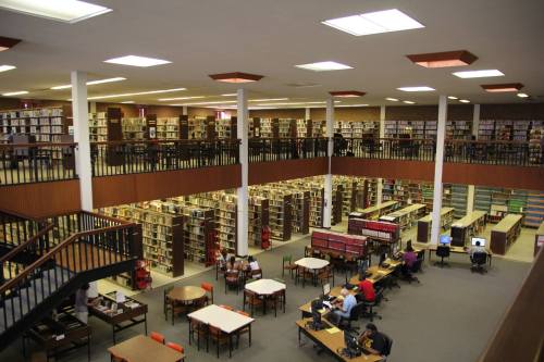 A spacious library with multiple levels, filled with bookshelves and study areas, and people engaged in reading and working.