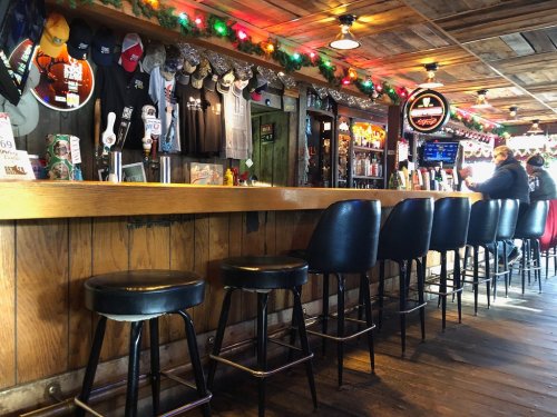 A cozy bar with a wooden counter, black stools, festive lights, and various hats and shirts displayed on the walls.