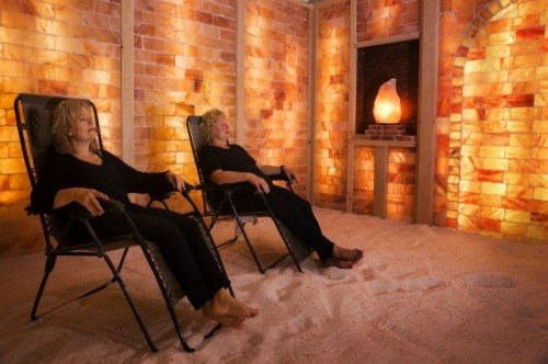 Two women relax in a salt therapy room, sitting in chairs with warm, glowing salt walls and a salt lamp in the background.