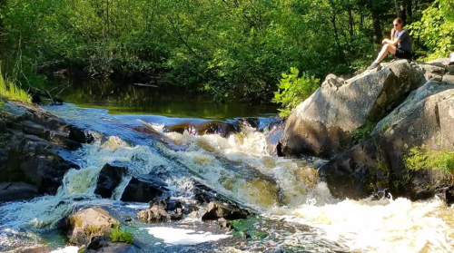 A person sits on a rock by a flowing river, surrounded by lush greenery and sunlight.