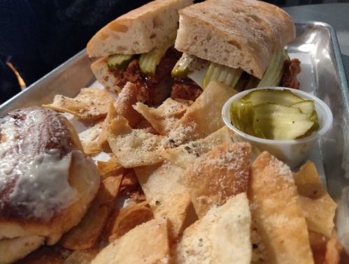 A plate with two sandwiches filled with shredded meat and pickles, served with crispy chips and a side of pickles.