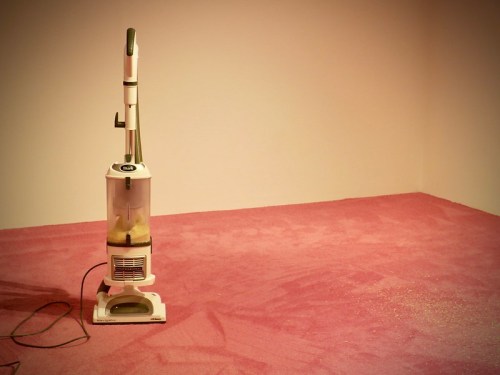 A white vacuum cleaner stands on a pink carpet, with visible cleaning marks and some scattered debris around it.