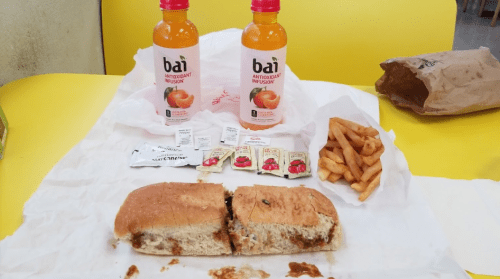 A split sandwich, two bottles of Bai drink, fries, and condiment packets on a yellow table.