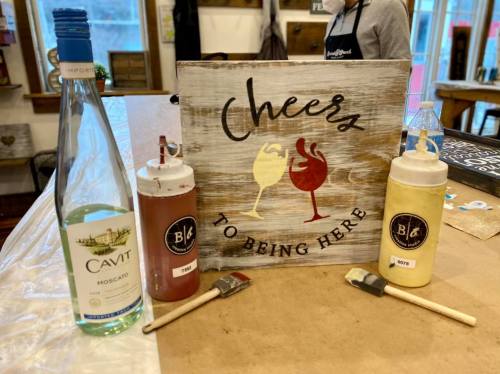 A wooden sign reading "Cheers to Being Here" with a bottle of wine and two condiment bottles on a table.