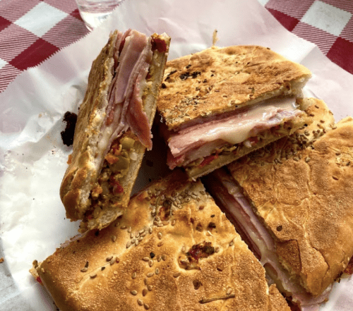 A close-up of a sliced sandwich with layers of ham, cheese, and colorful toppings on a toasted, sesame-seeded bread.