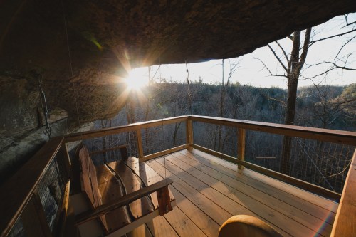 A wooden deck under a rock overhang, with a view of trees and a sunset in the background.
