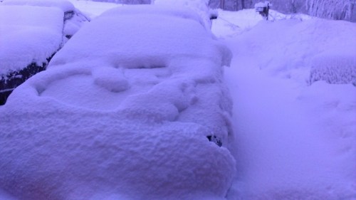 A car covered in thick snow, with a snowy landscape in the background. The scene appears cold and wintry.