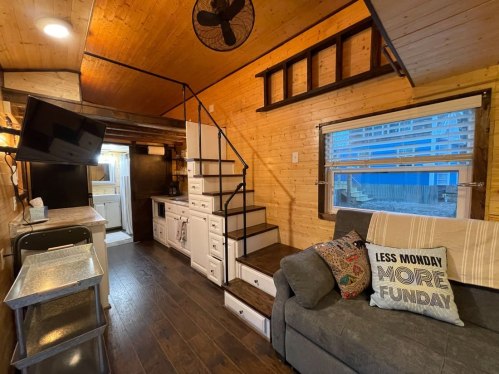 Cozy interior of a tiny home featuring a sofa, stairs, kitchen area, and wooden walls. Bright natural light from windows.