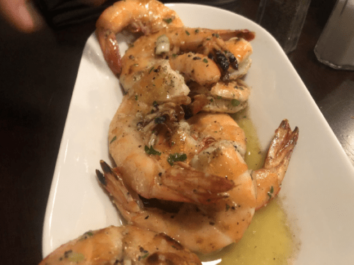 A white plate with several large, cooked shrimp drizzled with garlic butter and garnished with herbs.