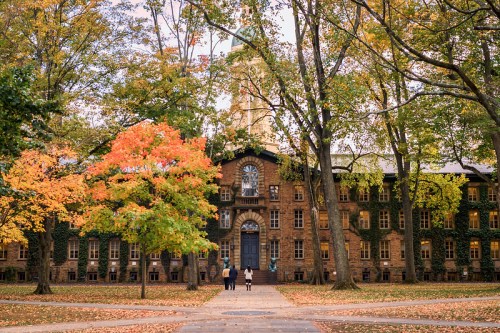 Two people walk towards a historic building surrounded by vibrant autumn trees and colorful foliage.
