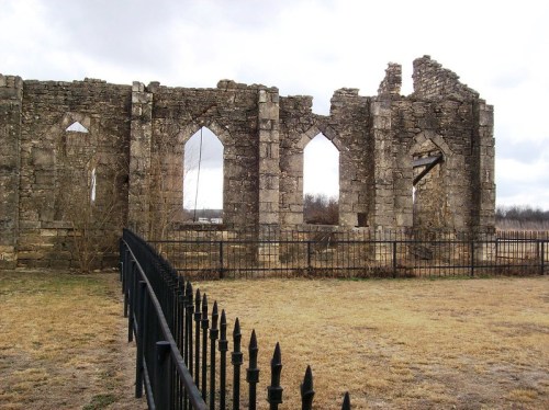 Ruins of a stone building with arched windows, surrounded by a black metal fence and grassy area under a cloudy sky.