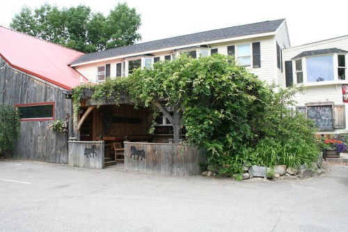 A rustic building with a wooden exterior, greenery, and a red roof, featuring outdoor seating and nearby flower beds.