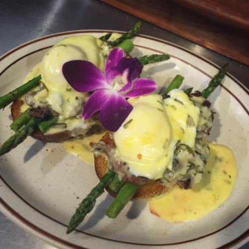 Two eggs Benedict on toasted bread with asparagus, topped with hollandaise sauce and a purple flower garnish.