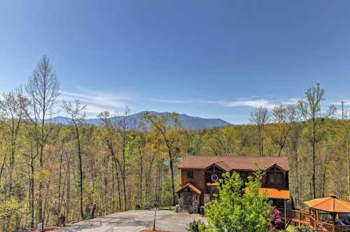 A scenic view of a mountain cabin surrounded by trees, with mountains visible in the background under a clear blue sky.