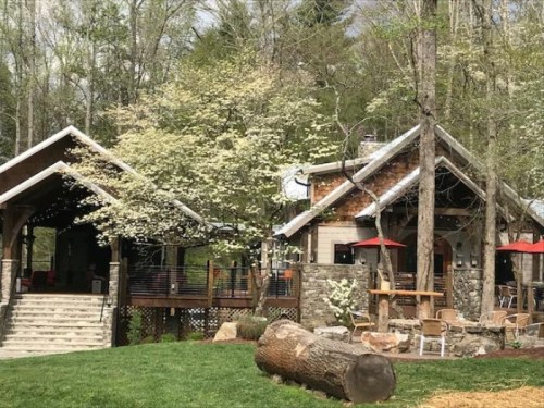 A rustic building surrounded by trees, featuring a stone facade, outdoor seating, and blooming flowers in the foreground.