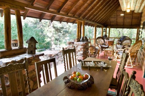 A rustic porch with wooden furniture, a dining table with fruit, and a view of greenery outside.