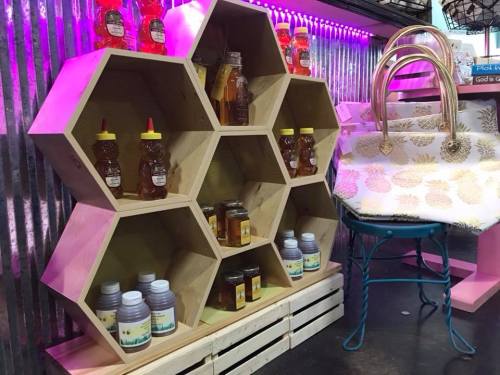 A hexagonal wooden display holds jars of honey and other products, alongside a stylish handbag on a blue chair.