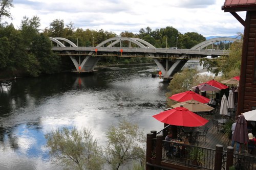 A modern bridge spans a river, with outdoor seating and umbrellas along the bank, under a cloudy sky.