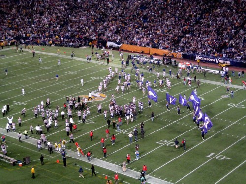 Aerial view of a football field with players and fans celebrating, flags waving, and a vibrant crowd in the stands.