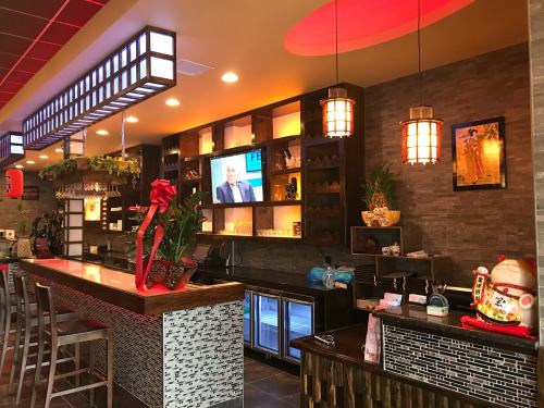 A modern restaurant interior featuring a bar, shelves with decor, and a TV on the wall, with warm lighting and plants.