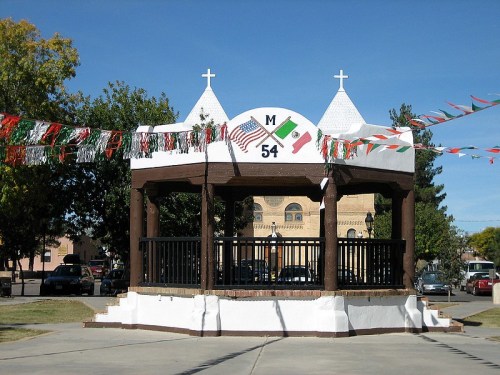 A decorated gazebo with flags and banners, set in a park with trees and a building in the background.