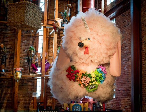 A large, fluffy pink poodle sculpture with a unicorn horn, adorned with colorful flowers, hanging in a rustic space.
