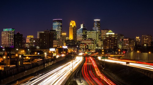 Minneapolis skyline at night, featuring illuminated buildings and light trails from moving traffic.