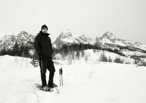 A person in winter gear stands on snow with mountains in the background, holding snowshoes.
