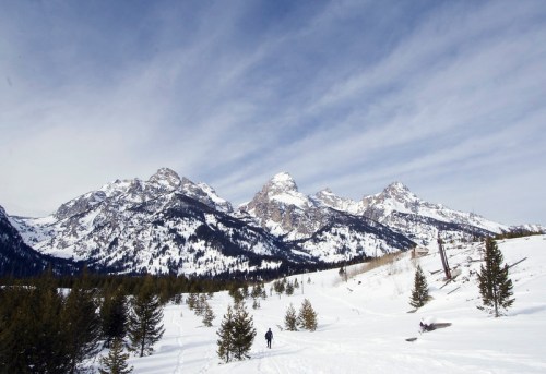 A person walks through a snowy landscape with towering mountains and a blue sky in the background.